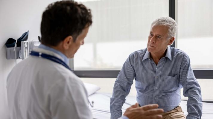 A patient sits on an exam table and listens to the doctor in the foreground talking