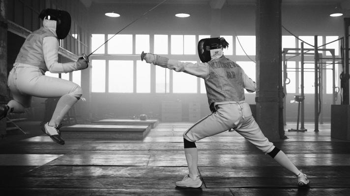 Black and white image of two individuals fencing. One is jumping but the opponent finds the weak spot. 