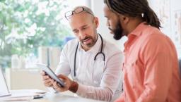 A male doctor sits with another man and they are looking at SDOH data on a mobile device.